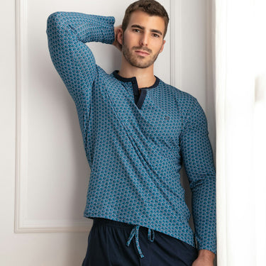 Man wearing a blue patterned shirt and dark pants against a white wall.